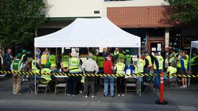 People gathered, many wearing bright safety vests, around a white tent behind a caution tape barrier at an outdoor event.