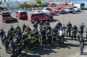Group of firefighters standing in formation with fire department vehicles in the background.